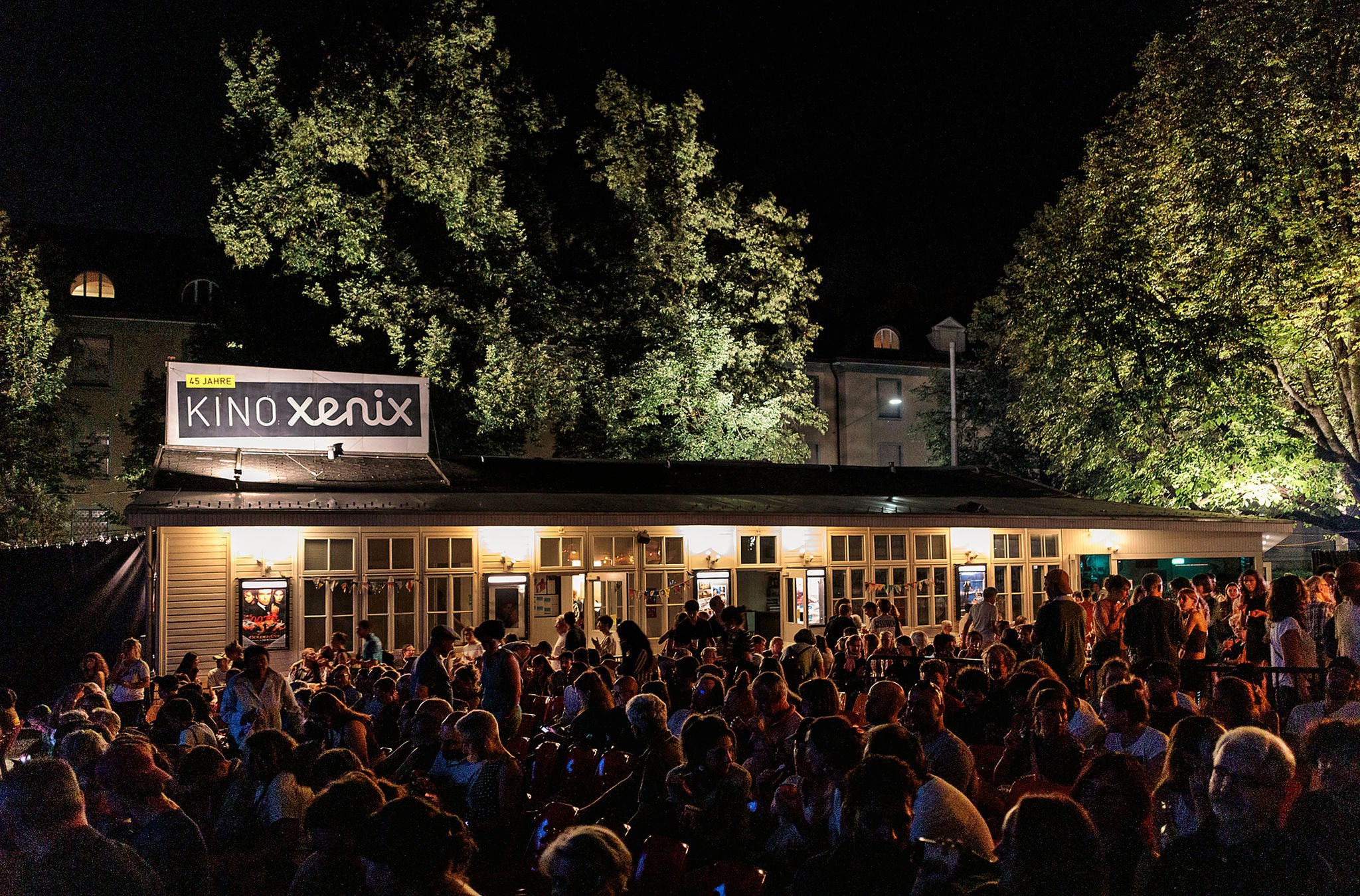 Der Aussenbereich des Xenix Kino und Bar Zürich hell beleuchtet  mit vielen Gästen bei Nacht.