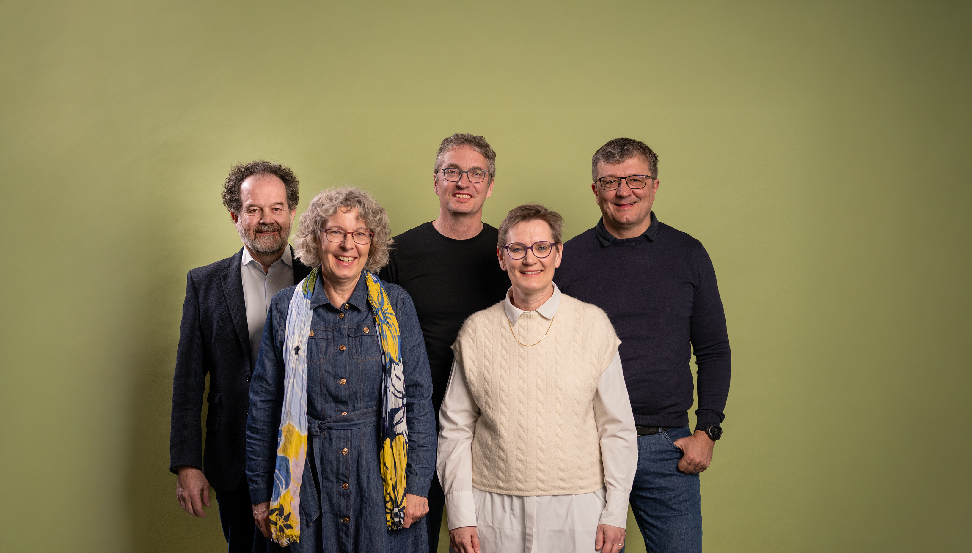 Eine Gruppe von fünf Erwachsenen steht vor einem hellgrünen Hintergrund und lächelt in die Kamera. Zwei Frauen stehen vorne, eine trägt ein langes Jeanskleid mit buntem Schal, die andere ein weisses Hemd mit cremefarbener Strickweste. Dahinter stehen drei Männer, alle mit Brille; sie tragen legere bis halbformelle Kleidung. Die Stimmung ist freundlich und entspannt.
