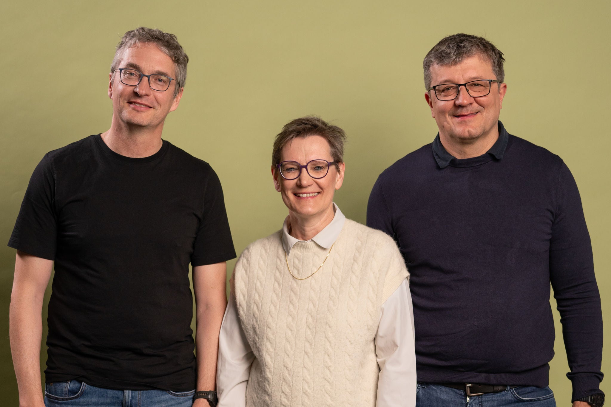 Drei Erwachsene stehen nebeneinander vor einem hellgrünen Hintergrund und lächeln in die Kamera. In der Mitte steht eine Frau mit kurzem Haar, Brille, weissem Hemd und cremefarbener Strickweste. Links von ihr steht ein Mann mit grauem Haar, Brille, schwarzem T-Shirt und Jeans. Rechts steht ein weiterer Mann mit Brille, dunkelblauem Pullover und Jeans. Alle wirken freundlich und entspannt.
