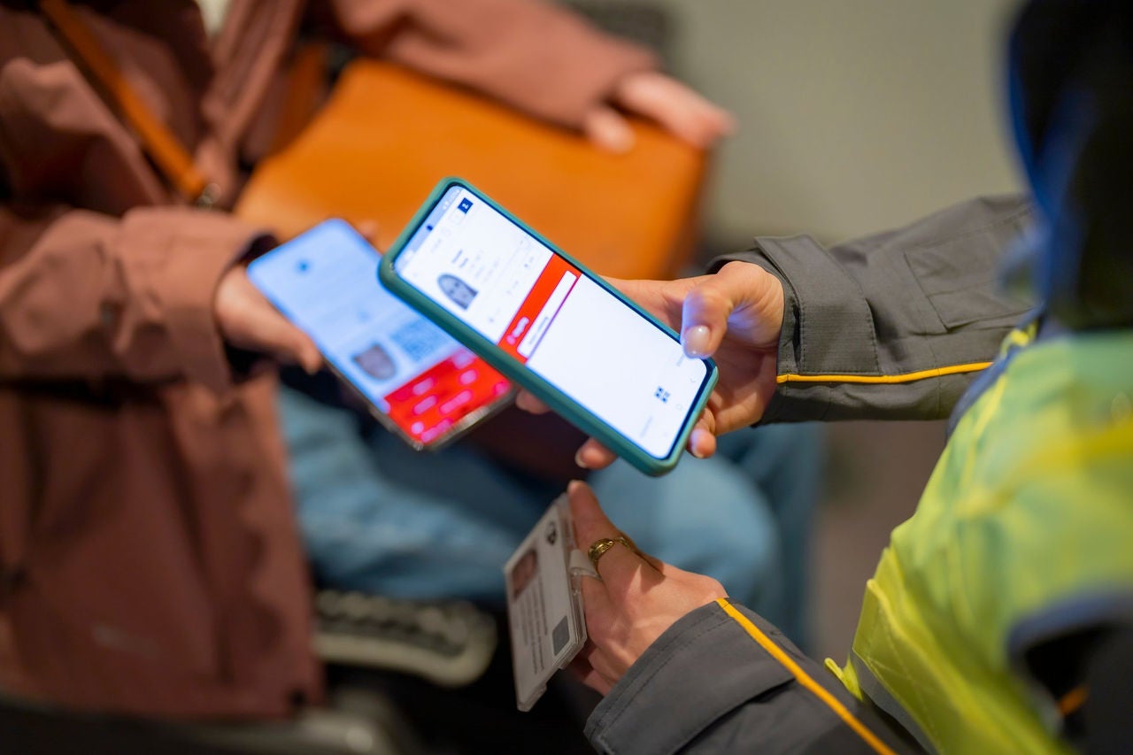 Nahaufnahme einer Hand, die ein Smartphone hält. Auf dem Display ist wird der SwissPass einer Person wird angezeigt. Am Bildrand ist erkennbar, dass die abgebildete Person eine gelbe Leuchtweste trägt. Im Hintergrund ist unscharf eine zweite, sitzende Person in einer braunen Jacke zu sehen zu sehen. Diese hat ebenfalls ein Smartphone in der Hand und zeigt diesen der ersten Person. Das Bild zeigt eine Ticketkontrolle im öffentlichen Verkehr. 