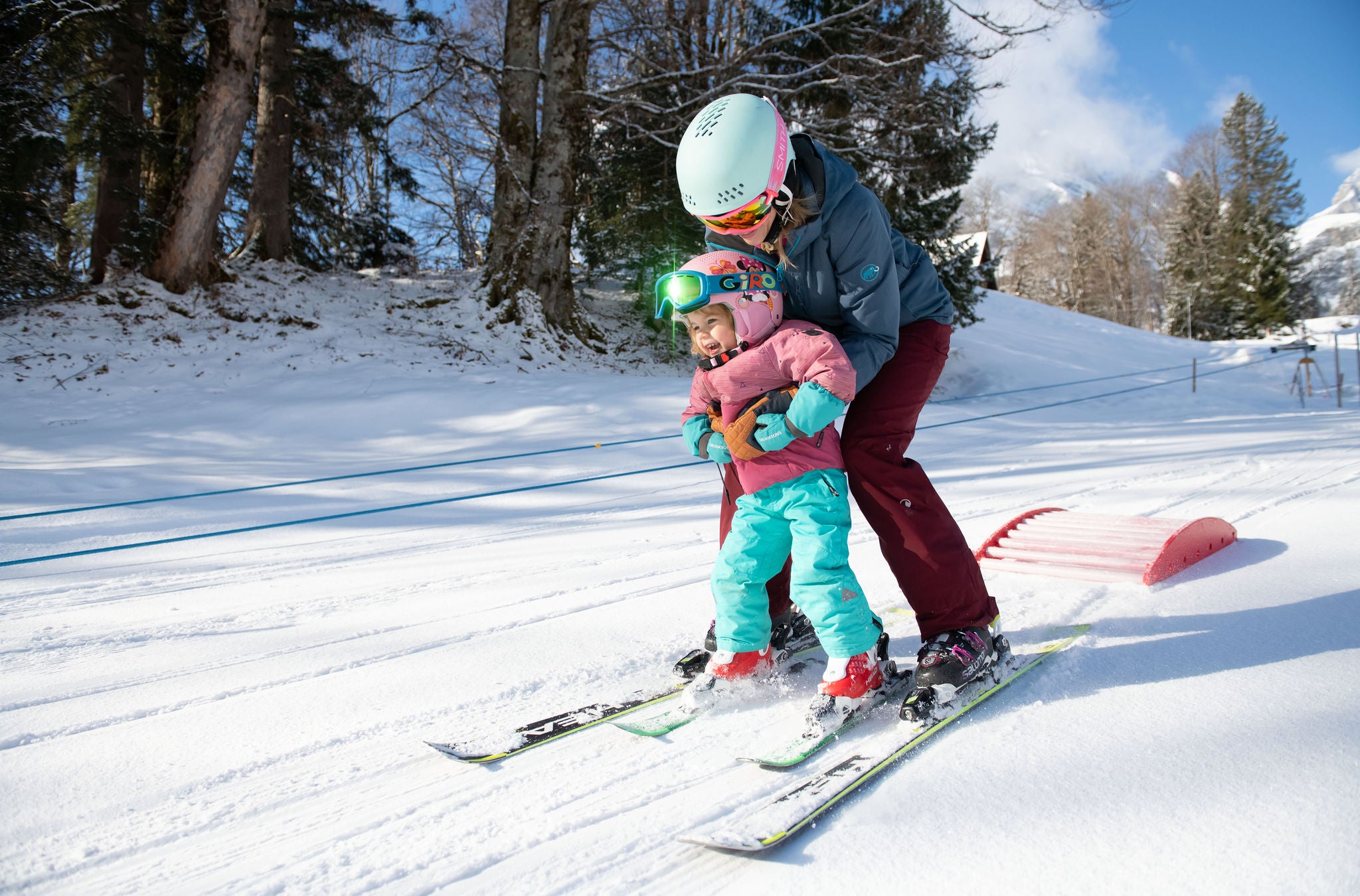 Eine erwachsene Person hält ein Kind aufrecht auf den Skis. 