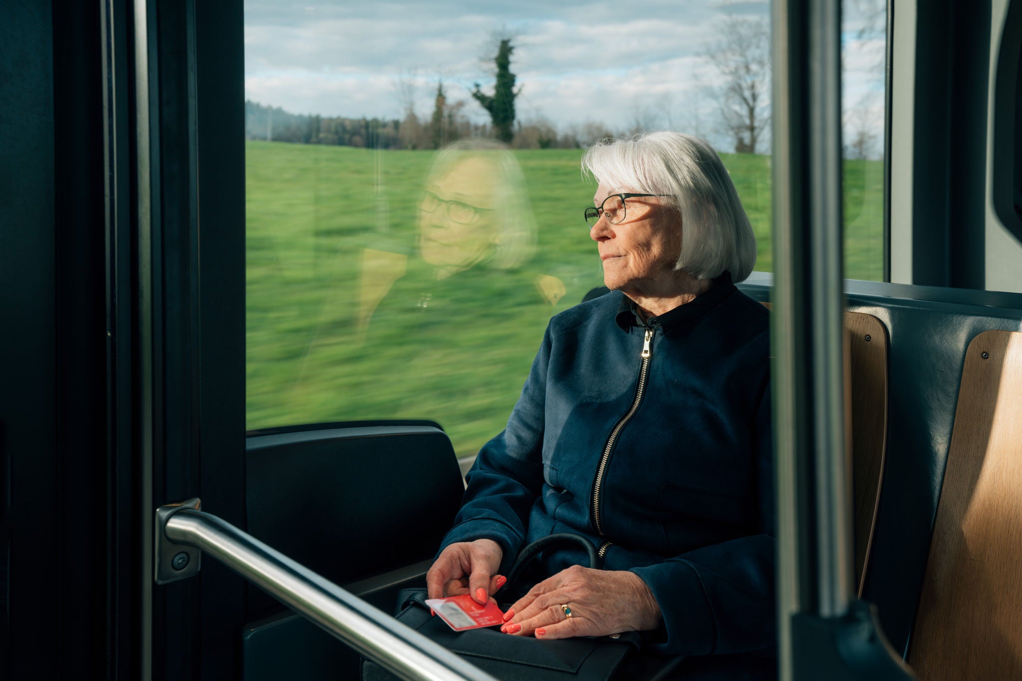 Seniorin sitzt im öV schaut aus dem Fenster und hält einen Swisspass in dern Händen