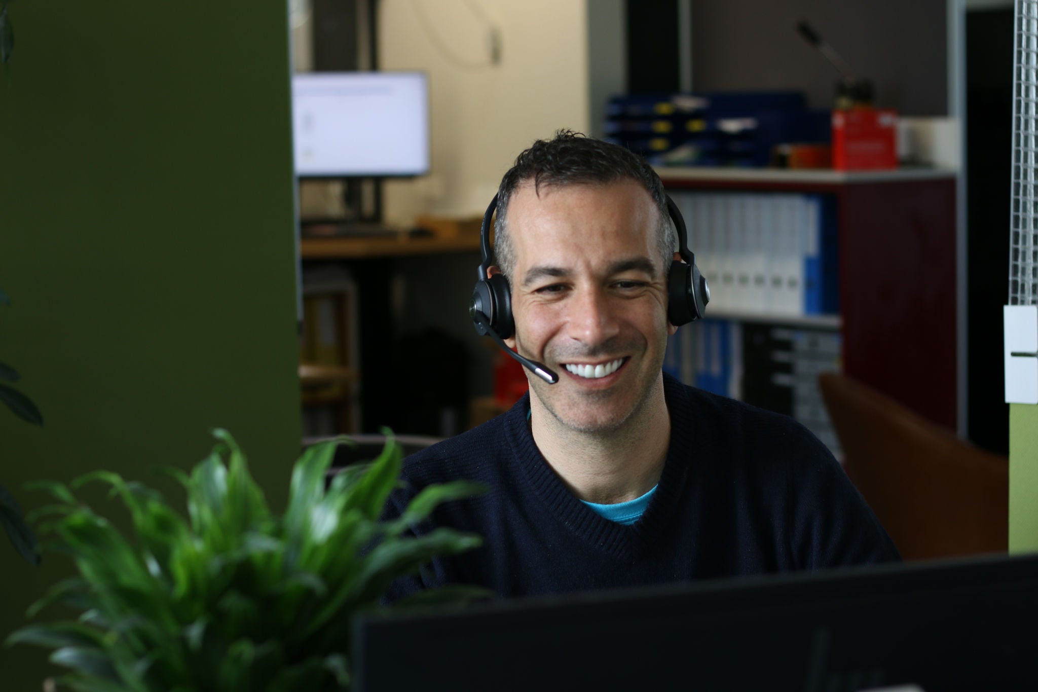 Ein lächelnder Mann mit Headset sitzt an einem Schreibtisch im Büro und schaut auf einen Bildschirm, im Hintergrund sind Ordner und ein Computer zu sehen.