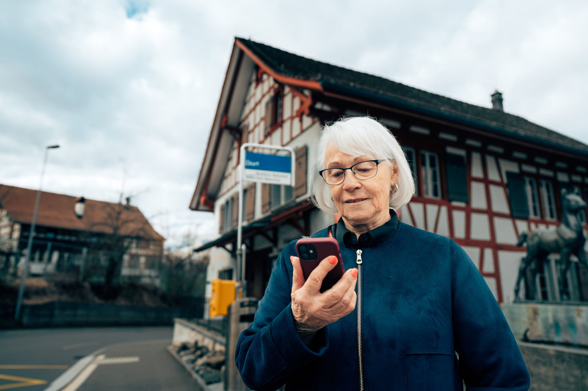 Frau mit weissen Haaren steht vor Bushaltestelle und schaut auf ihr Smartphone