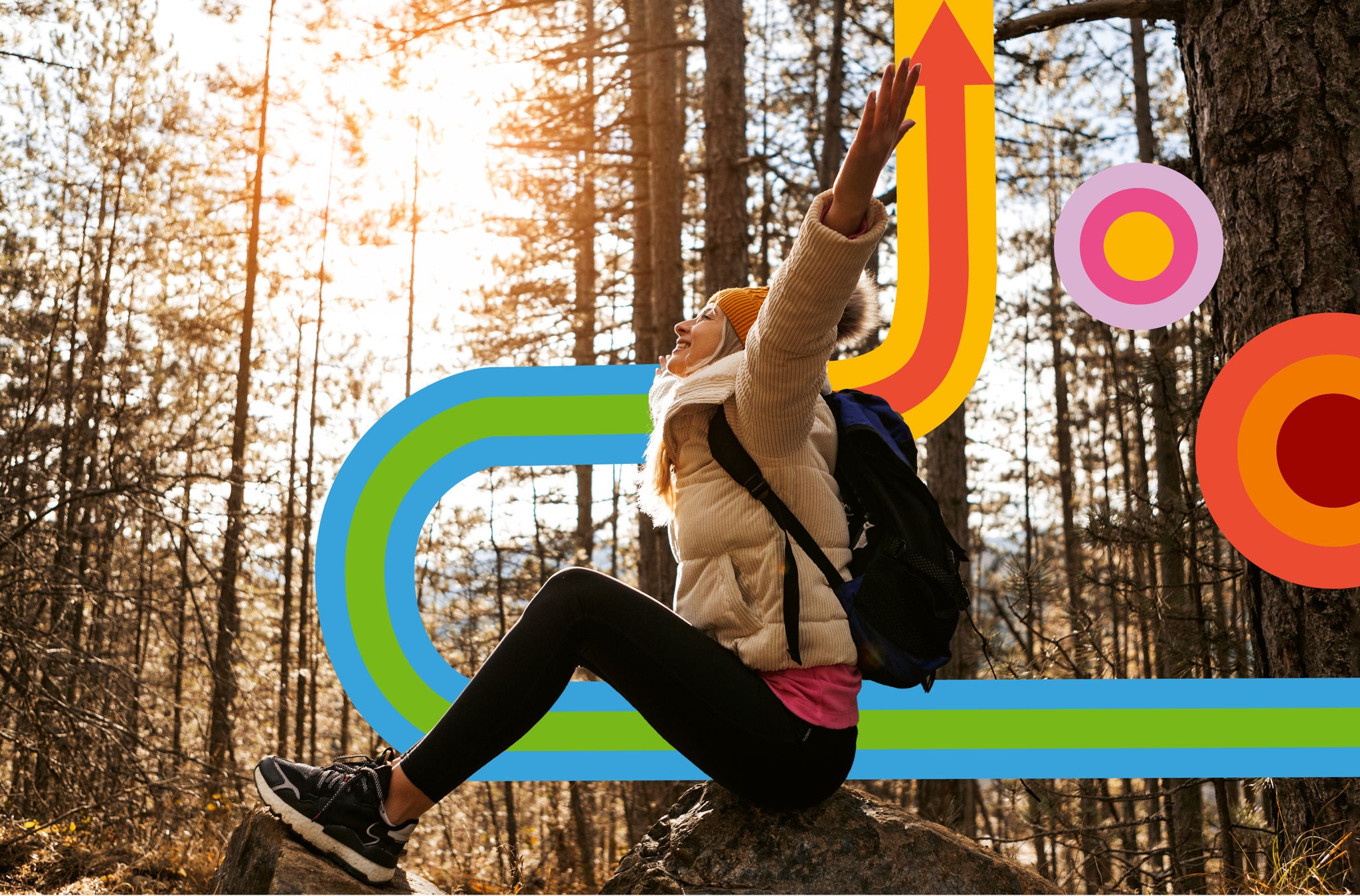 Frau in Outdoor-Kleidern sitzt im Wald auf einem Stein und hält die Hände in die Luft.