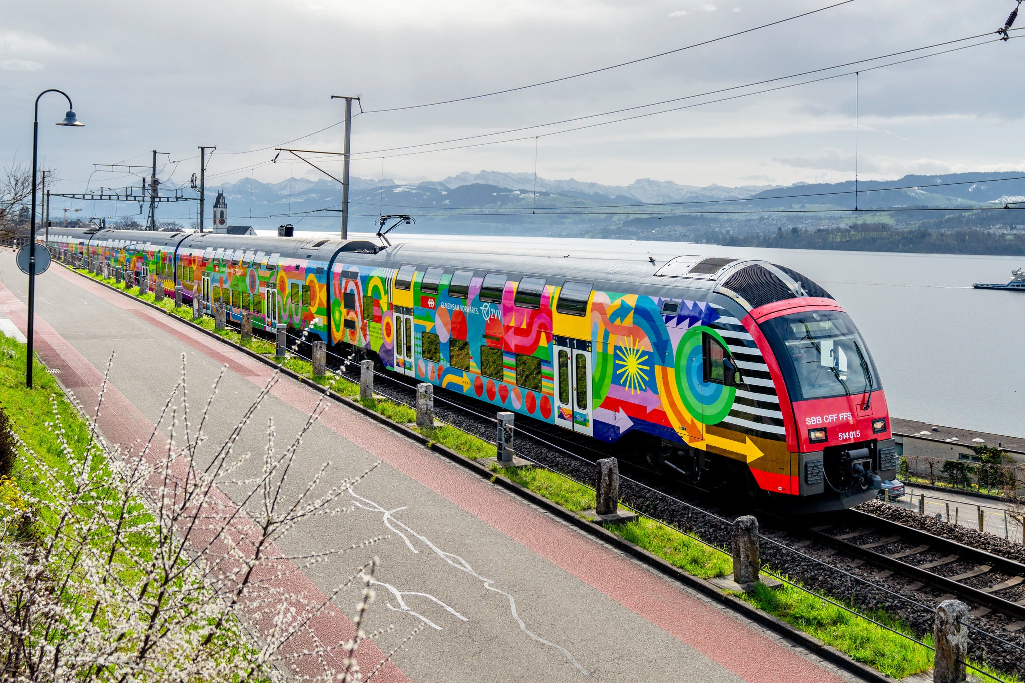 Ein farbenfroh bemalter Doppelstockzug der SBB (Zugnummer 514 015) fährt entlang eines Sees (Zürichsee) mit schneebedeckten Alpen im Hintergrund. Die auffällige Bemalung zeigt abstrakte Muster (ZVV Kampagne Gemeinsam Vorwärts)und leuchtende Farben. Der Zug fährt auf Gleisen parallel zu einem Rad- und Fussweg, im Vordergrund sind blühende Zweige zu sehen
