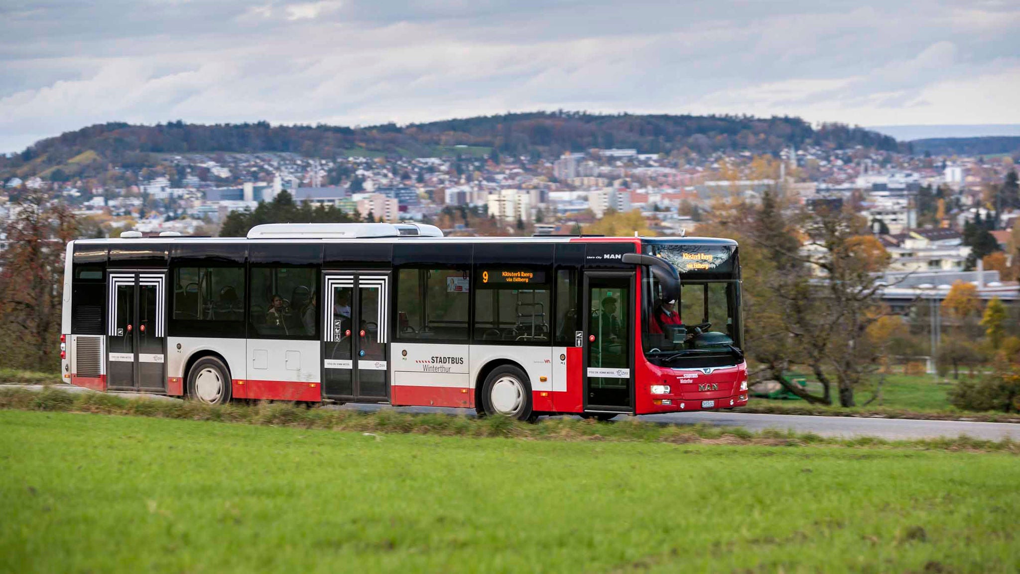 Weiss-roter Bus von Stadtbus Winterthur auf einer Landstrasse. Im Vordergrund eine Wiese, im Hintergrund eine Stadt.