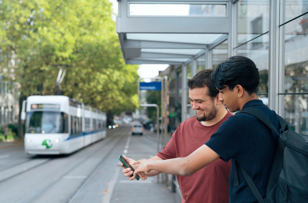 Zwei Männer stehen an einer Tramhaltestelle und schauen gemeinsam aufs Smartphone.
