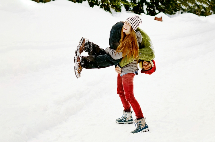 Eine Frau trägt lachend ihren Freund auf der Schulter. Sie sind in einer verschneiten Umgebung. 