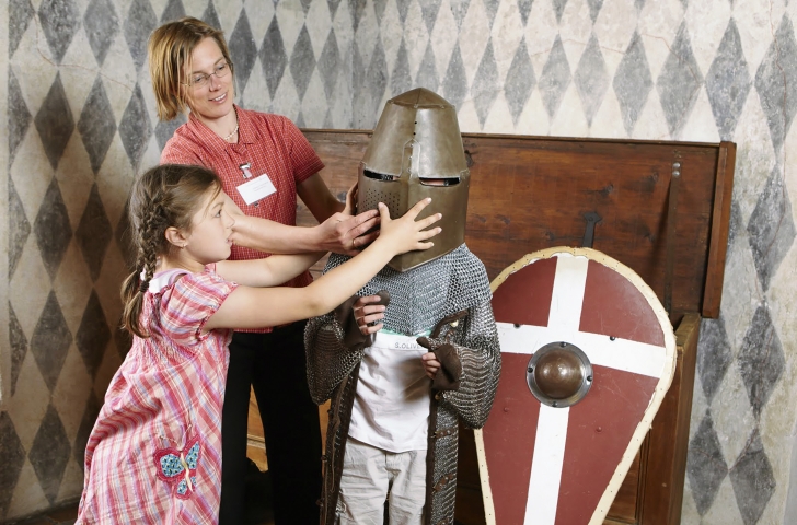 Frau und Mädchen helfen einem Jungen im Ritterhaus Bubikon eine Ritterausrüstung anzuziehen.
